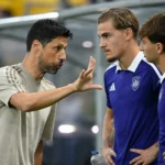Een coach geeft aanwijzingen aan spelers in paarse shirts langs de zijlijn.