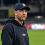 Een coach in donkere hoodie en pet naast de zijlijn na een wedstrijd.