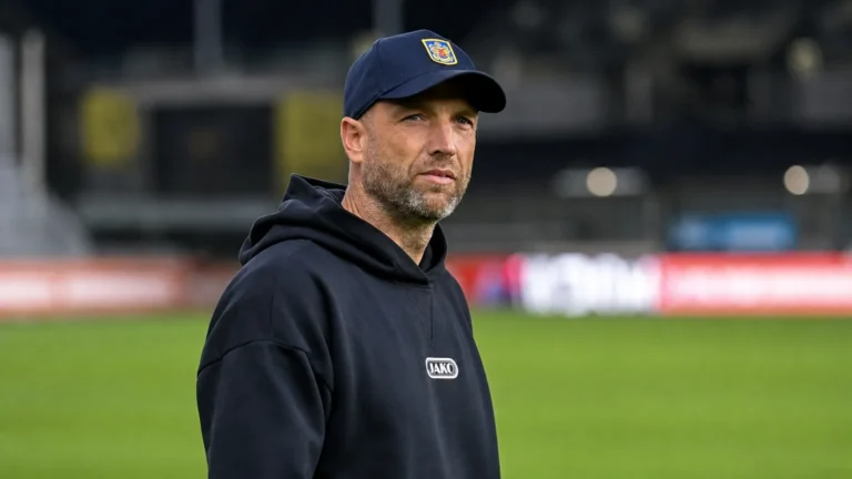 Een coach in donkere hoodie en pet naast de zijlijn na een wedstrijd.