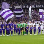 Voetballers en jeugdmascottes tijdens een gezamenlijke opstelling voor een wedstrijd, met paarse supportersvlaggen op de tribune.