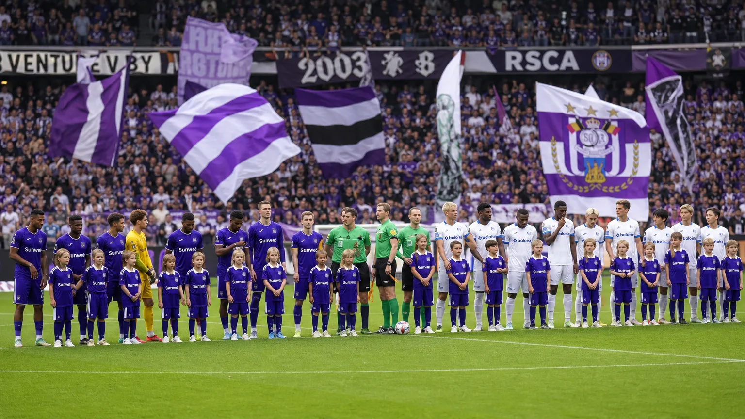 Voetballers en jeugdmascottes tijdens een gezamenlijke opstelling voor een wedstrijd, met paarse supportersvlaggen op de tribune.
