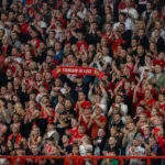Een volle groep Standard-supporters in rood-witte kledij op de tribune met een Standard-sjaal.