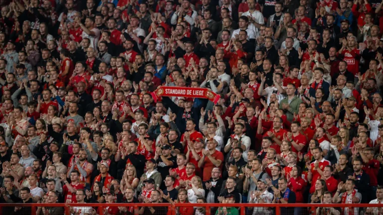 Een volle groep Standard-supporters in rood-witte kledij op de tribune met een Standard-sjaal.