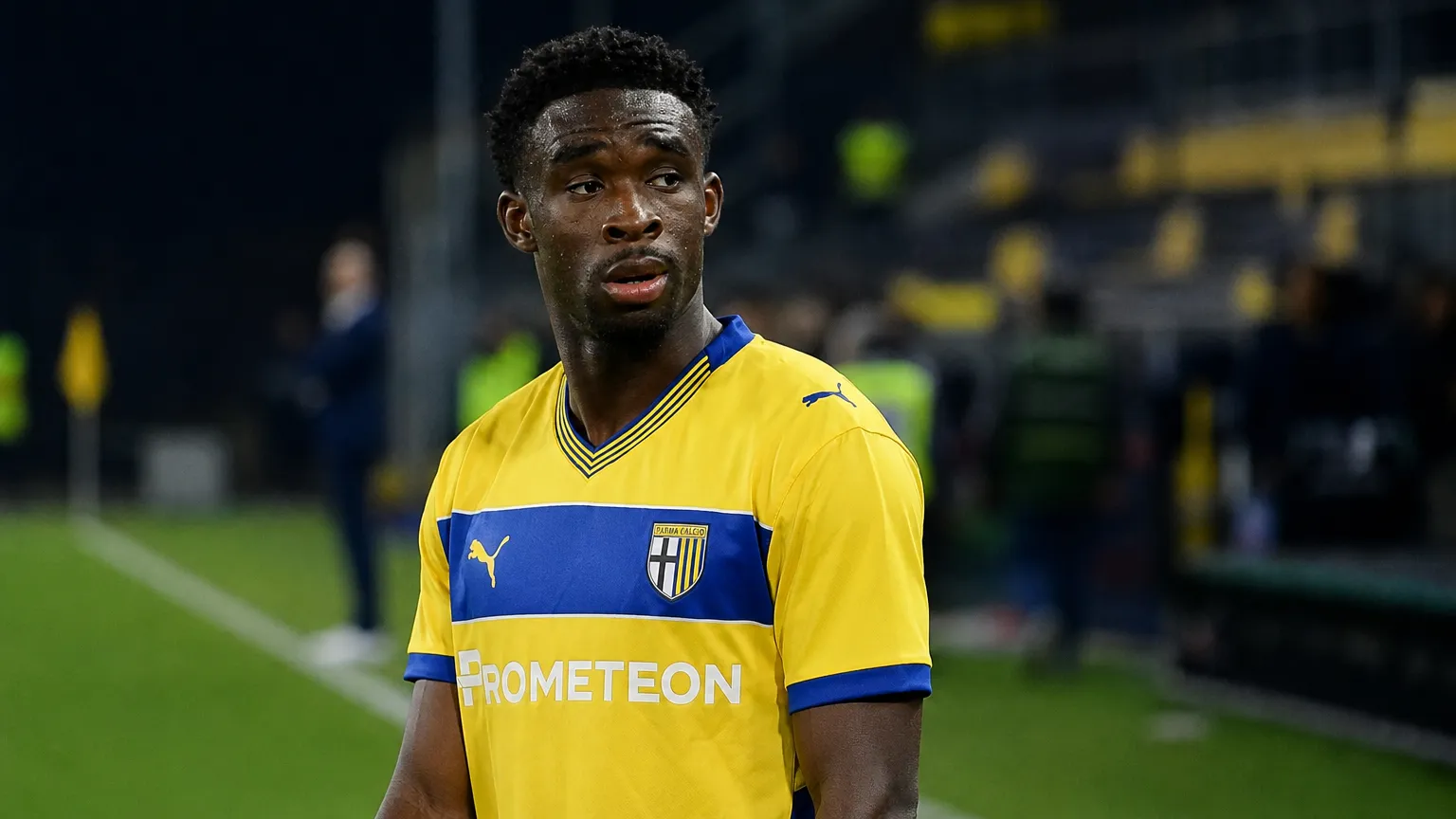 Mandela Keita in geel-blauwe spelerskleding langs de zijlijn van een voetbalveld.