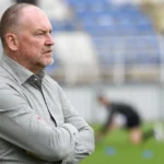Fred Vanderbiest kijkt geconcentreerd toe vanaf de zijlijn van een voetbalstadion.