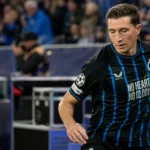 Een voetballer in een zwart-blauw gestreept tenue warmt op naast het veld in een gevuld stadion.