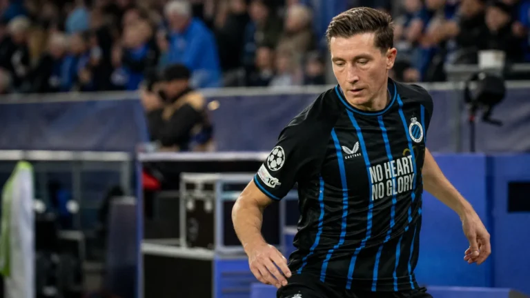 Een voetballer in een zwart-blauw gestreept tenue warmt op naast het veld in een gevuld stadion.