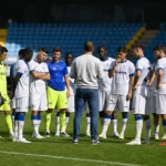 Een spelersgroep in witte tenues verzameld op het voetbalveld.