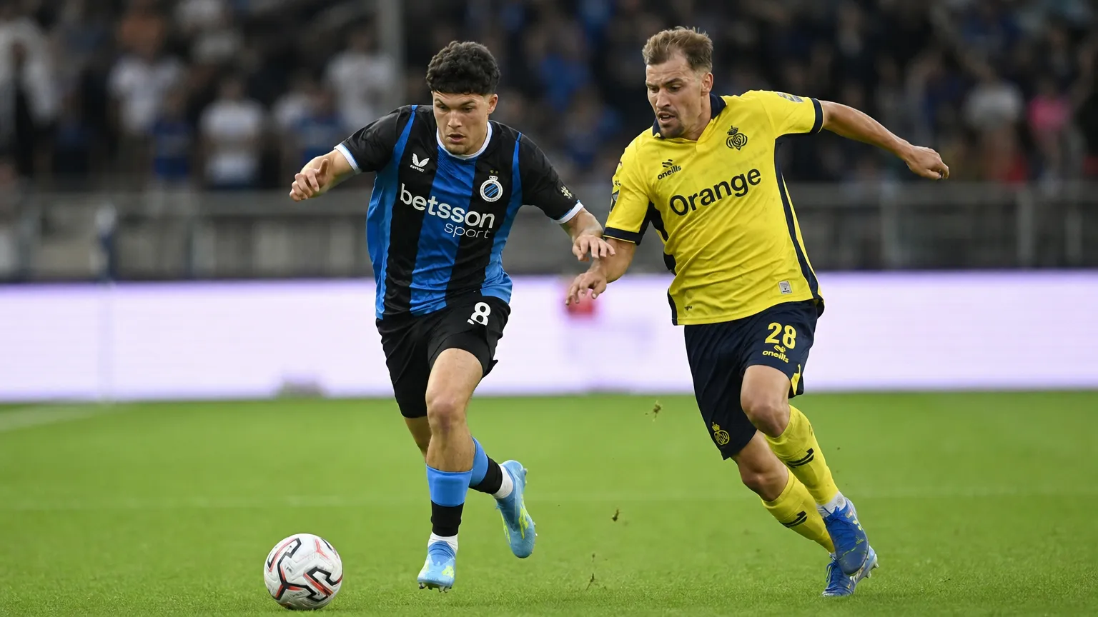 Twee voetballers strijden om de bal op een Belgisch voetbalveld.