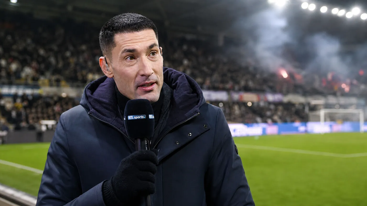 Silvio Proto spreekt tijdens een voetbalinterview in een donkere jas in een stadionomgeving.