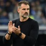 Een coach in zwarte trui applaudisseert langs de rand van een voetbalveld.