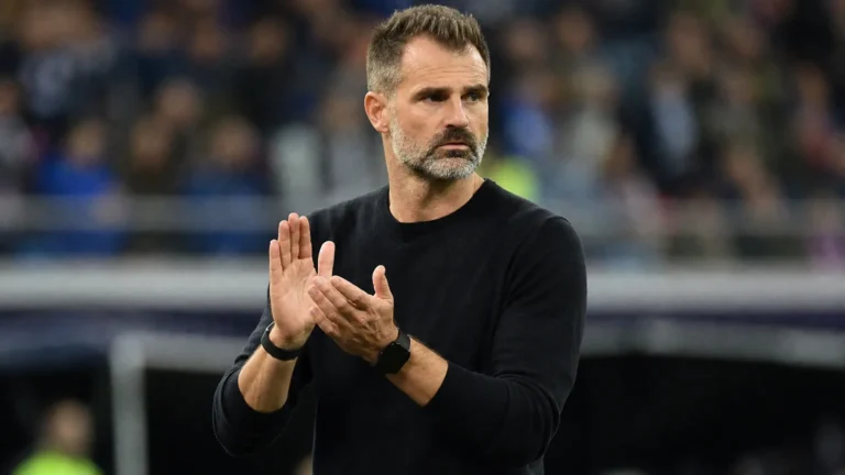 Een coach in zwarte trui applaudisseert langs de rand van een voetbalveld.