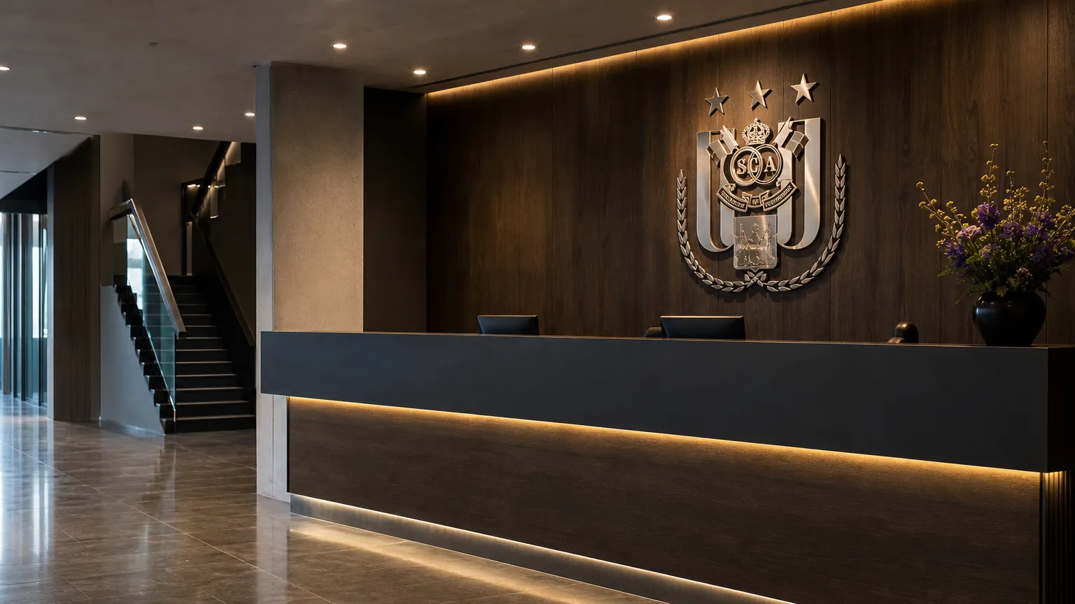 De lege verlichte inkomhal van het RSC Anderlecht-clubgebouw met een vast embleem aan de wand.