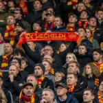 Een dicht opeengepakte groep KV Mechelen-supporters met rood-gele sjaals op de thuistribune.