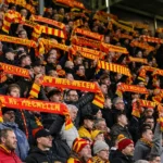 Supporters in rood-geel heffen sjaals in de tribune van een voetbalstadion.