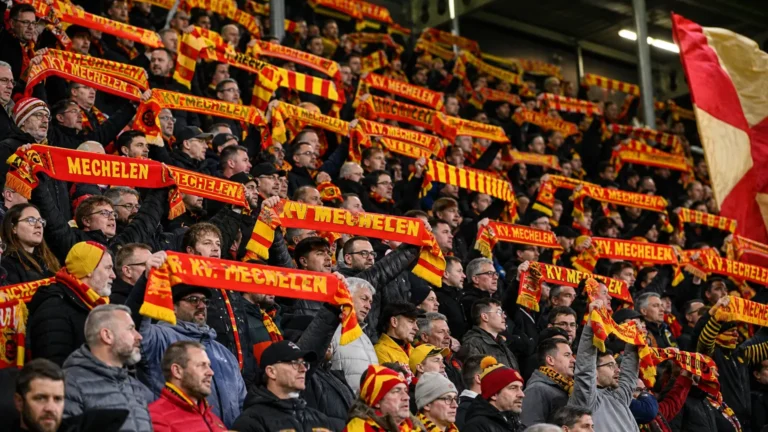 Supporters in rood-geel heffen sjaals in de tribune van een voetbalstadion.