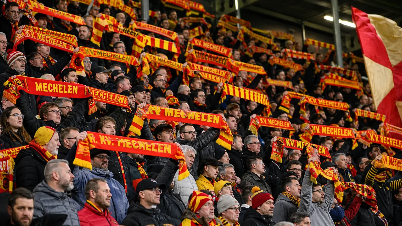 Supporters in rood-geel heffen sjaals in de tribune van een voetbalstadion.