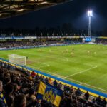 Het compacte ’t Kuipje van KVC Westerlo met het speelveld, overdekte tribunes en geel-blauwe accenten in een Kempense voetbalsfeer.