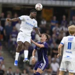 Spelers in witte en paarse tenues vechten een luchtduel uit tijdens een voetbalwedstrijd.
