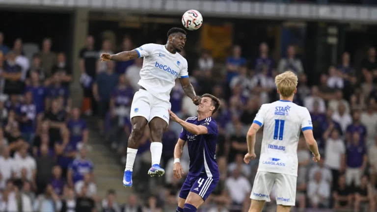 Spelers in witte en paarse tenues vechten een luchtduel uit tijdens een voetbalwedstrijd.