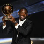 Een voetballer in een donker pak houdt de Ballon d'Or-trofee vast op een prijzenpodium.