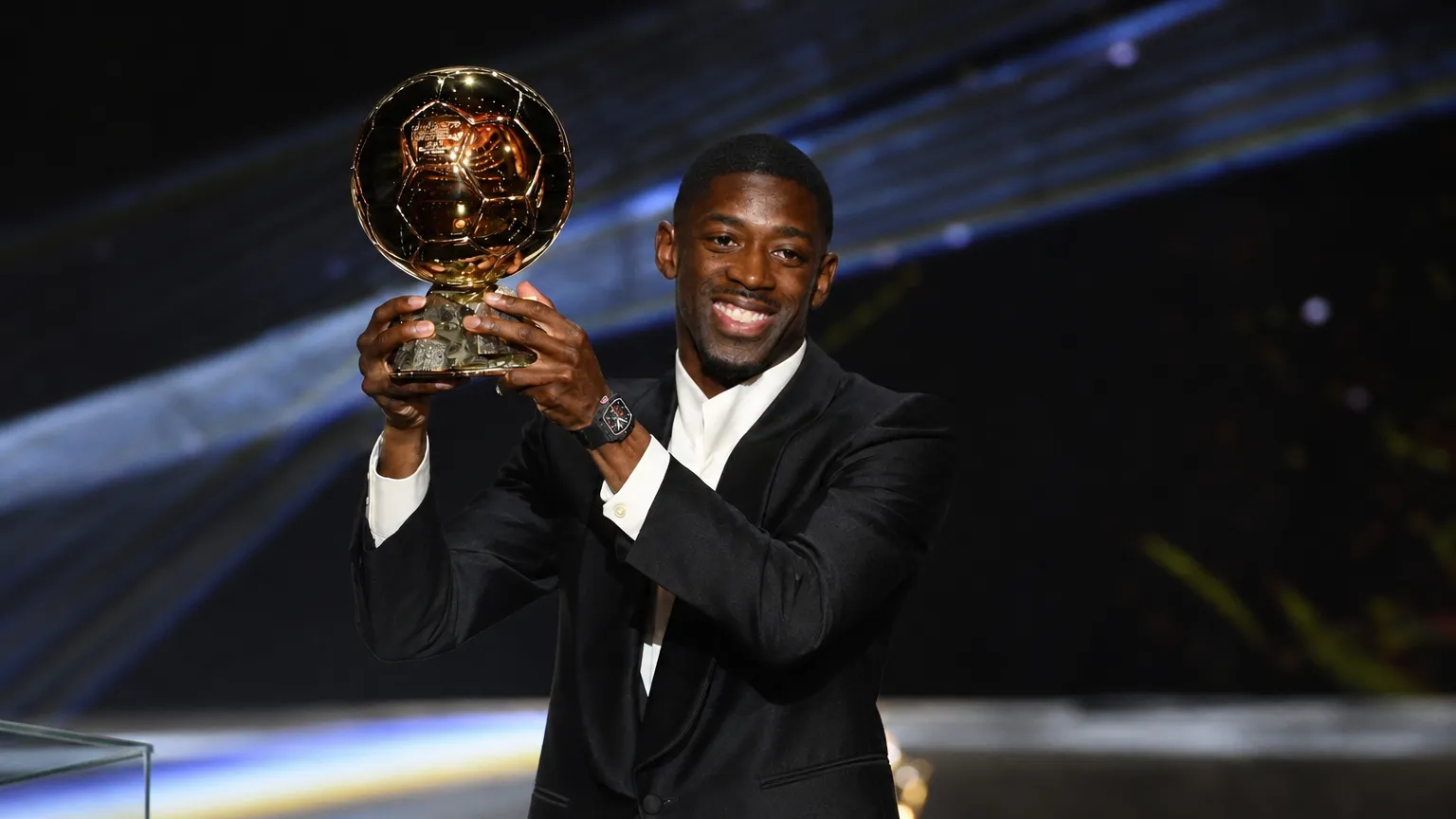 Een voetballer in een donker pak houdt de Ballon d'Or-trofee vast op een prijzenpodium.