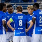 Een groep KRC Genk-spelers in blauwe shirts verzamelt zich rustig langs het veld.