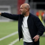 Een coach in donkere jas geeft aanwijzingen langs de zijlijn tijdens een voetbalwedstrijd.