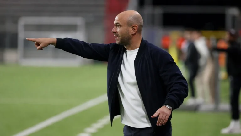 Een coach in donkere jas geeft aanwijzingen langs de zijlijn tijdens een voetbalwedstrijd.
