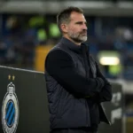 Een Club Brugge-trainer staat na de wedstrijd rustig langs de zijlijn bij een stadionbord.