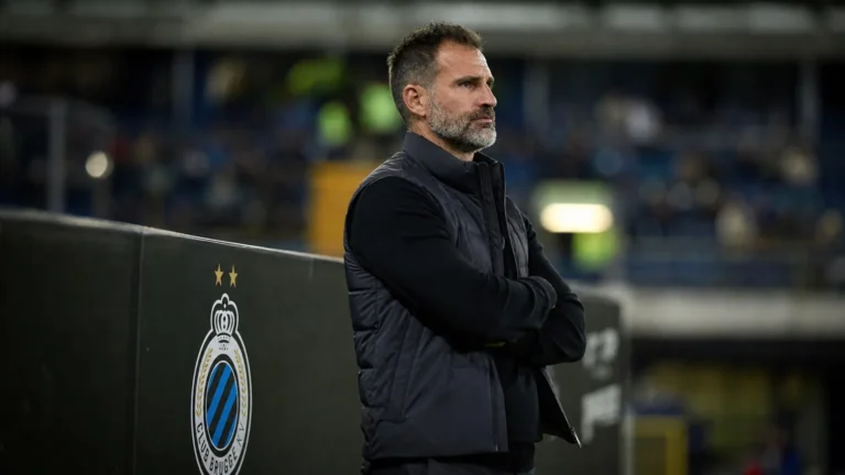 Een Club Brugge-trainer staat na de wedstrijd rustig langs de zijlijn bij een stadionbord.