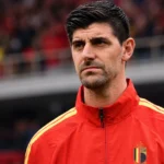 Thibaut Courtois in een rode trainingsjas van België aan de zijlijn van een voetbalveld.