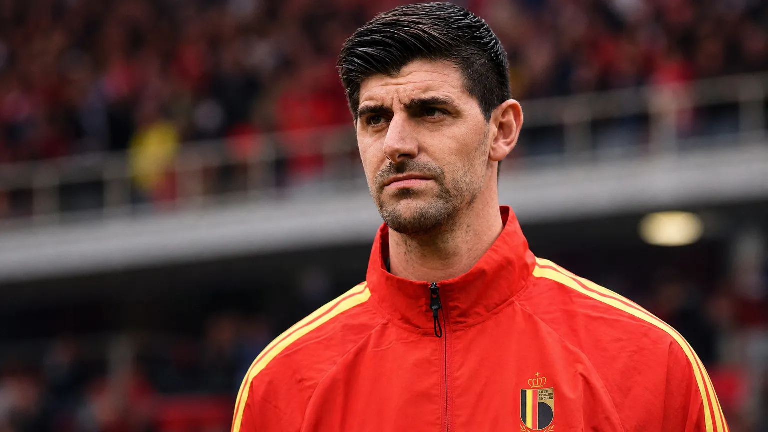 Thibaut Courtois in een rode trainingsjas van België aan de zijlijn van een voetbalveld.