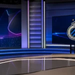 Lege televisiestudio voor een Champions League-voorbeschouwing met een Club Brugge-embleem op een vaste monitor.
