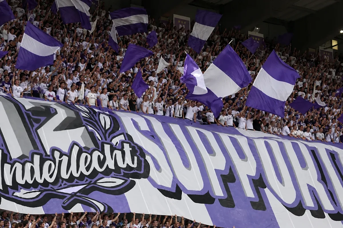 Een volle tribune met supporters die paarse en witte vlaggen zwaaien.