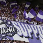 Een volle tribune met supporters die paarse en witte vlaggen zwaaien.