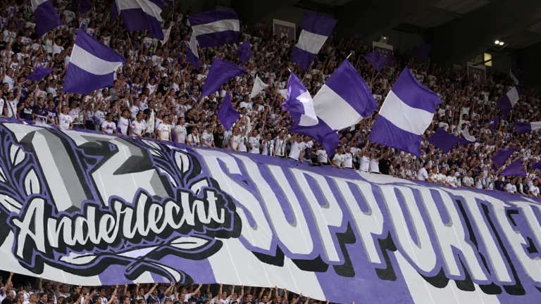 Een volle tribune met supporters die paarse en witte vlaggen zwaaien.