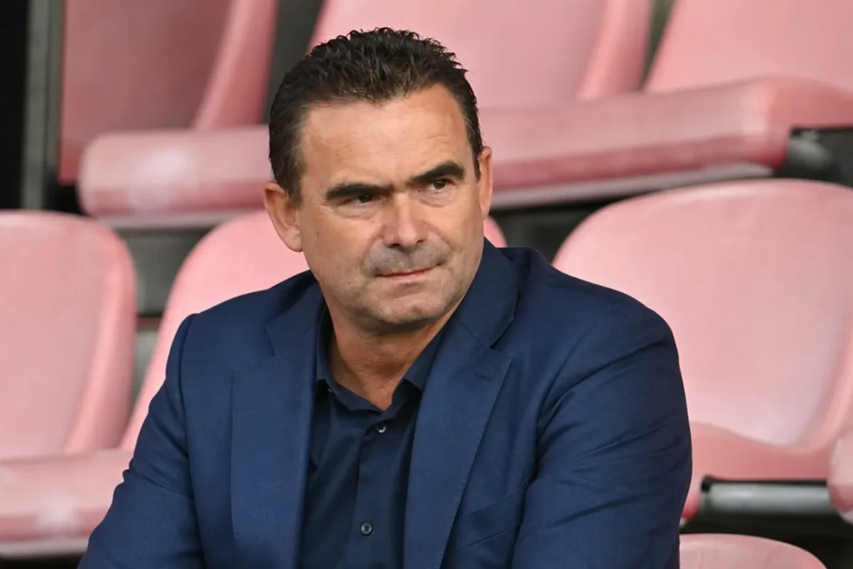 Marc Overmars zit in een rustige stadiontribune in een donkerblauw kostuum.