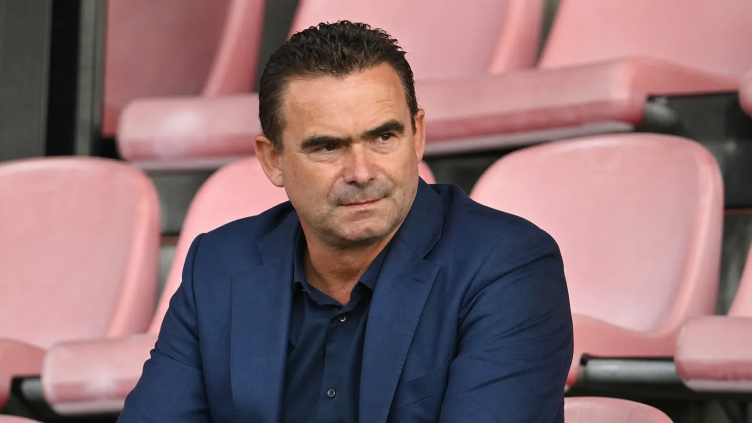 Marc Overmars zit in een rustige stadiontribune in een donkerblauw kostuum.