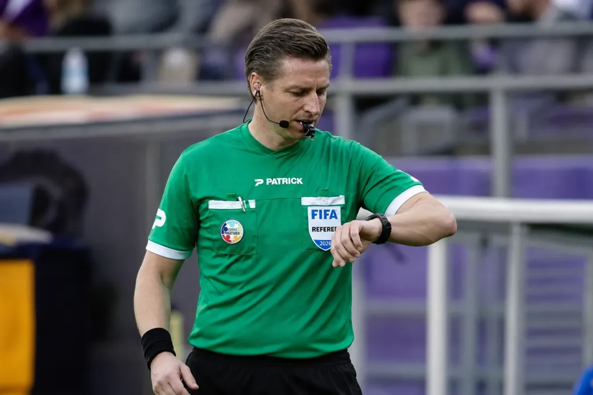 Een scheidsrechter in groen tenue staat voor de aftrap op een Belgisch voetbalveld.