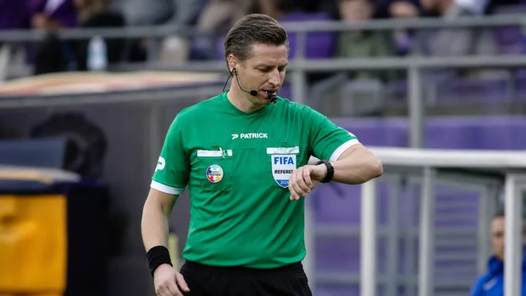 Een scheidsrechter in groen tenue staat voor de aftrap op een Belgisch voetbalveld.