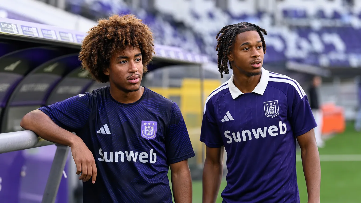 Twee jonge voetballers in paarse Anderlecht-tenues staan naast een voetbalveld.