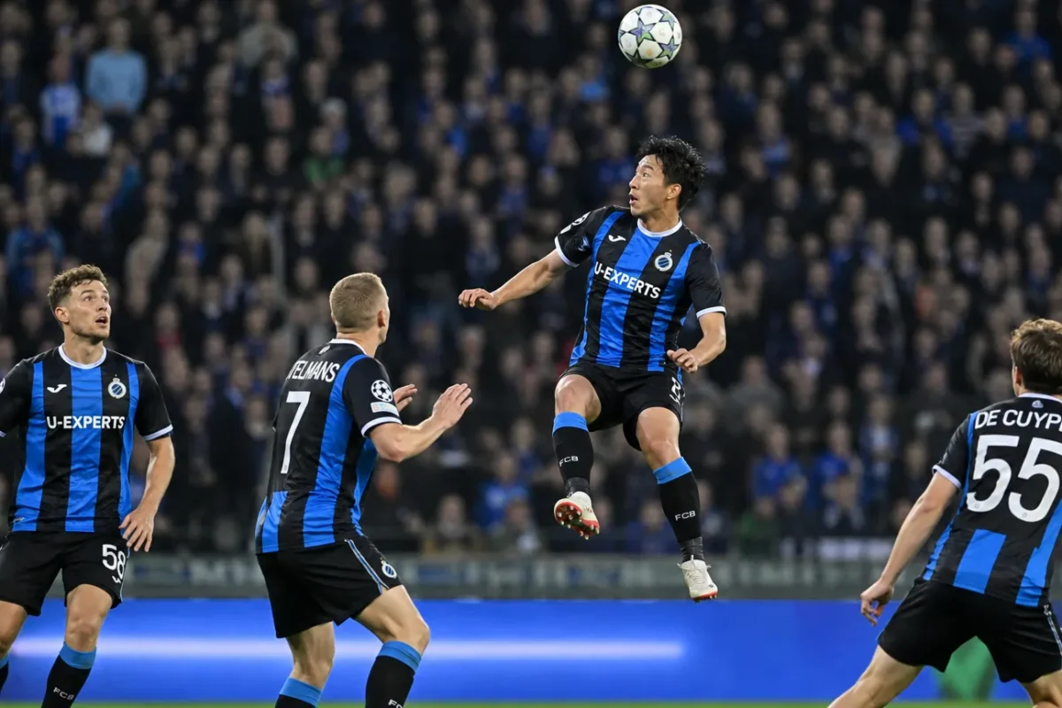 Voetballers in blauw-zwarte tenues vechten om een hoge bal in een avondwedstrijd.