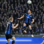 Voetballers in blauw-zwarte tenues vechten om een hoge bal in een avondwedstrijd.
