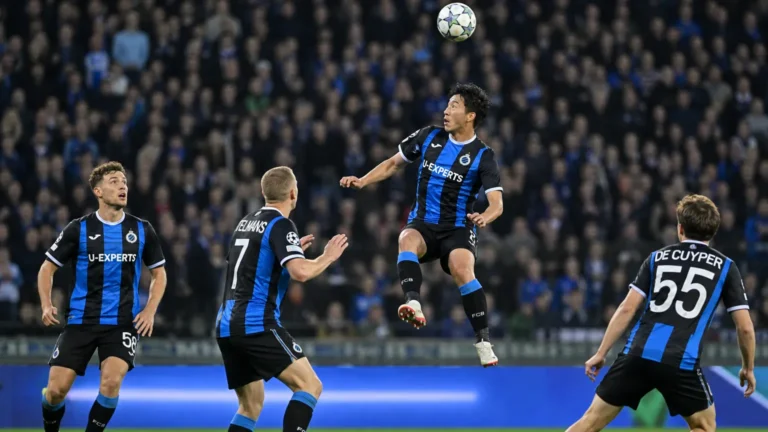 Voetballers in blauw-zwarte tenues vechten om een hoge bal in een avondwedstrijd.