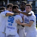 Een groep KAA Gent-spelers viert samen op het veld in witte shirts met blauwe accenten.
