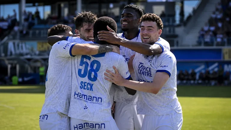 Een groep KAA Gent-spelers viert samen op het veld in witte shirts met blauwe accenten.