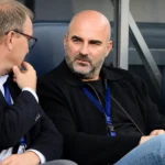 Twee niet-spelende clubvertegenwoordigers in gesprek op de bank van een voetbalstadion.