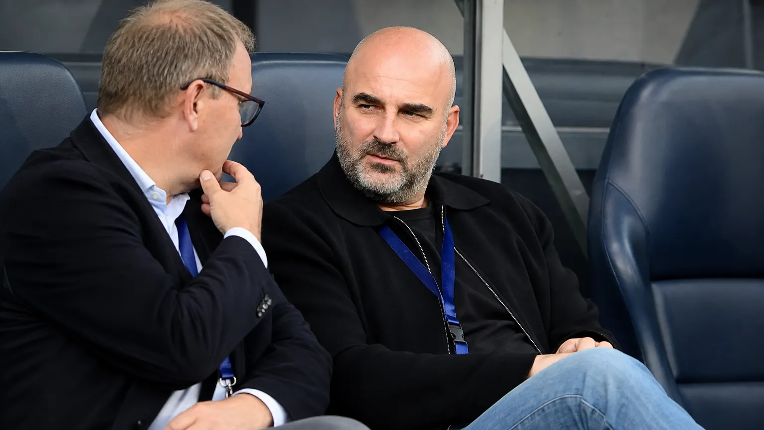 Twee niet-spelende clubvertegenwoordigers in gesprek op de bank van een voetbalstadion.