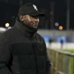 George Desmond Kamurasi aan de zijlijn van een Engels amateurvoetbalveld in donkere bovenkleding.